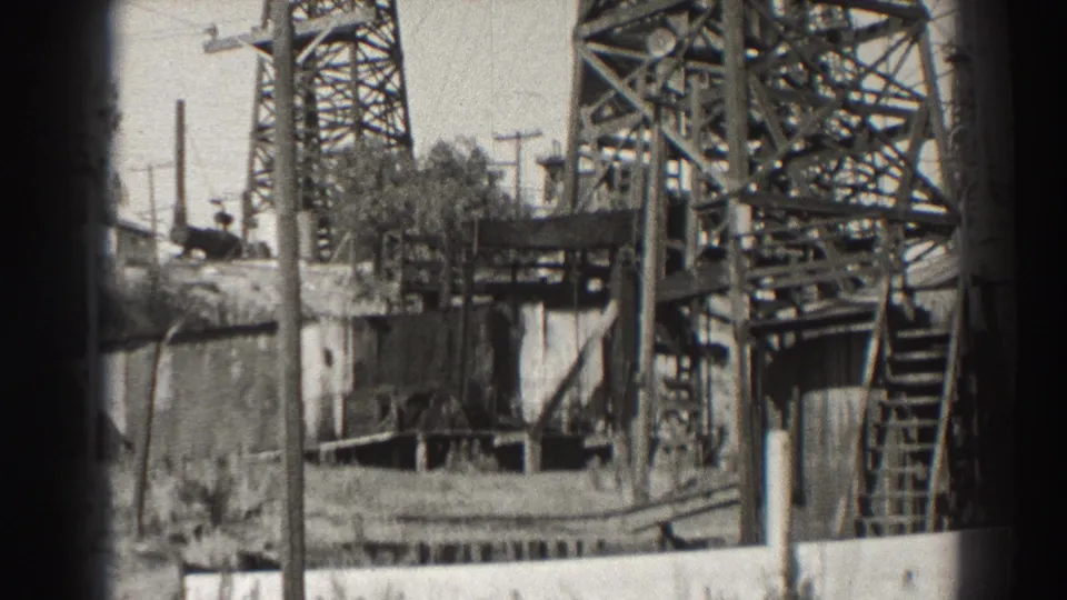 Black-and-white 1937 Long Beach frame of an oil well with lattice tower, laddered platform, small structure, poles, and brush.