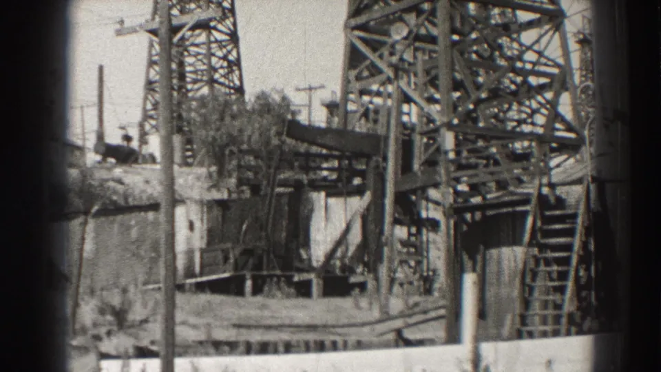 Black-and-white 8mm frame from Long Beach in 1937 showing an oil derrick, laddered platform, base building, poles, and vegetation.