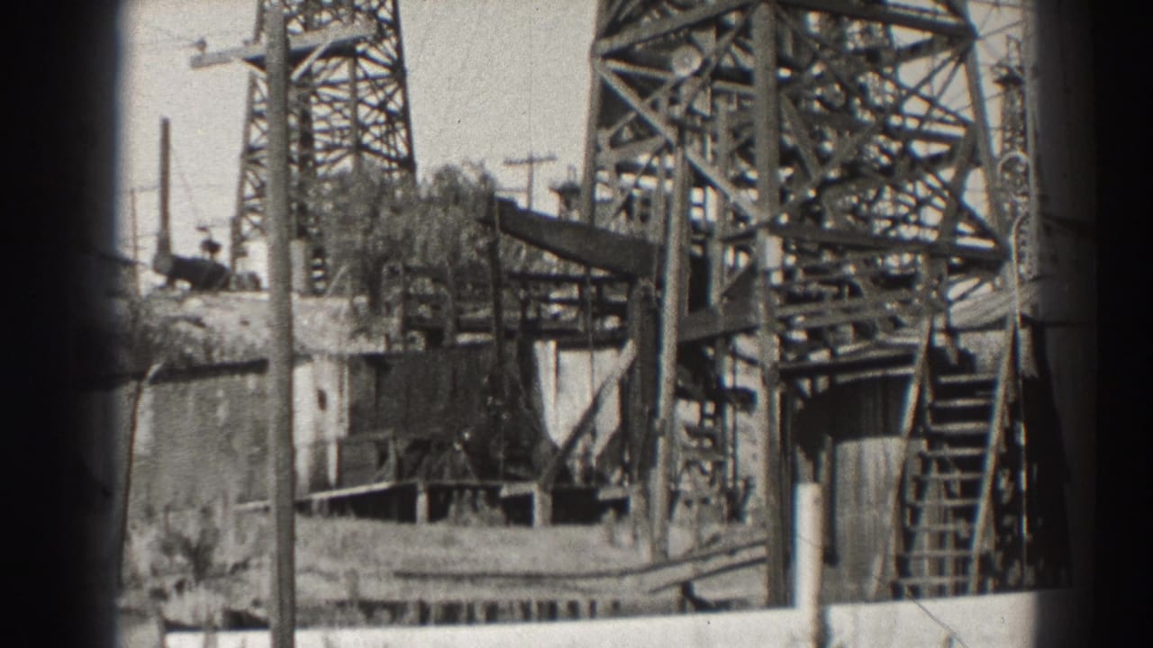 1937 Long Beach oil-field view with steel derrick framing, ladder, utility poles, wall, and extraction equipment.