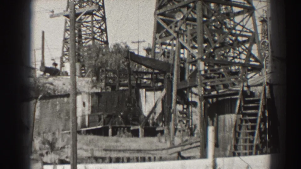 1937 Long Beach oil-field view with steel derrick framing, ladder, utility poles, wall, and extraction equipment.