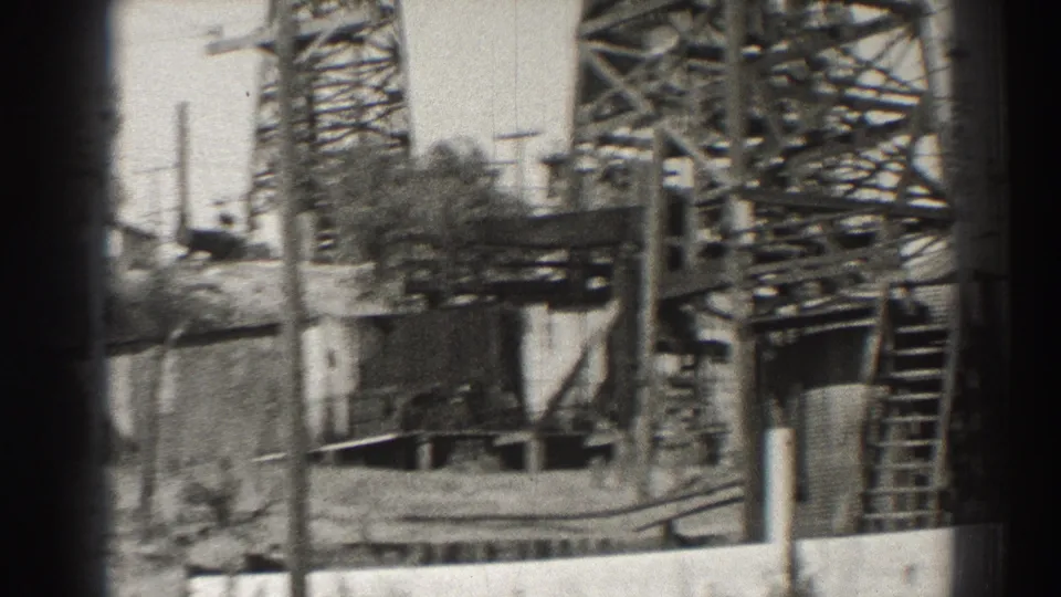 Long Beach oil-field machinery appears in soft focus, with derrick framing, ladder, wall, pole, and yard details.