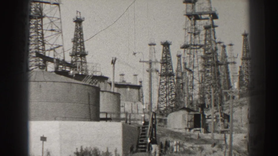 Wide 1937 Long Beach oil-field frame showing multiple derricks, storage tanks, stairs, utility poles, and overhead cables.