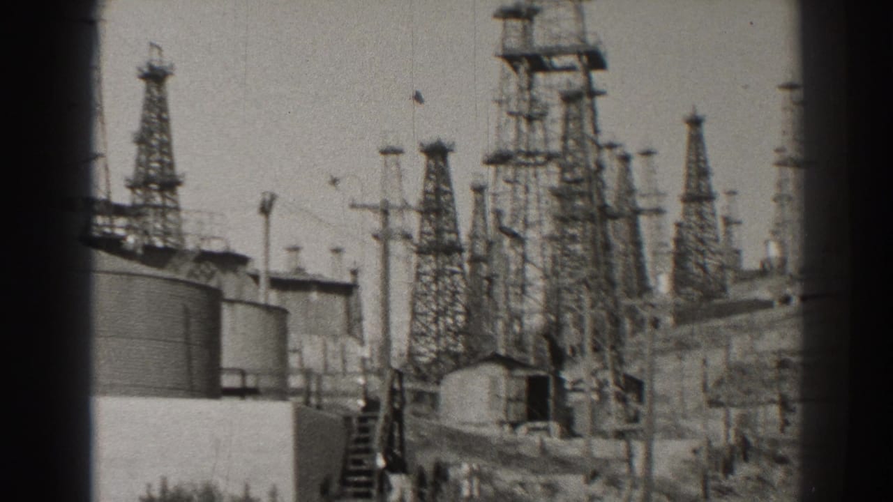 1937 Long Beach 8mm frame showing lattice transmission towers, cylindrical tanks, utility poles, and a small structure in a power-station yard.