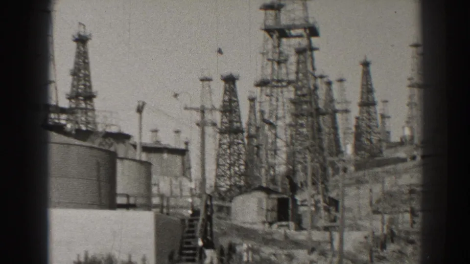 1937 Long Beach 8mm frame showing lattice transmission towers, cylindrical tanks, utility poles, and a small structure in a power-station yard.