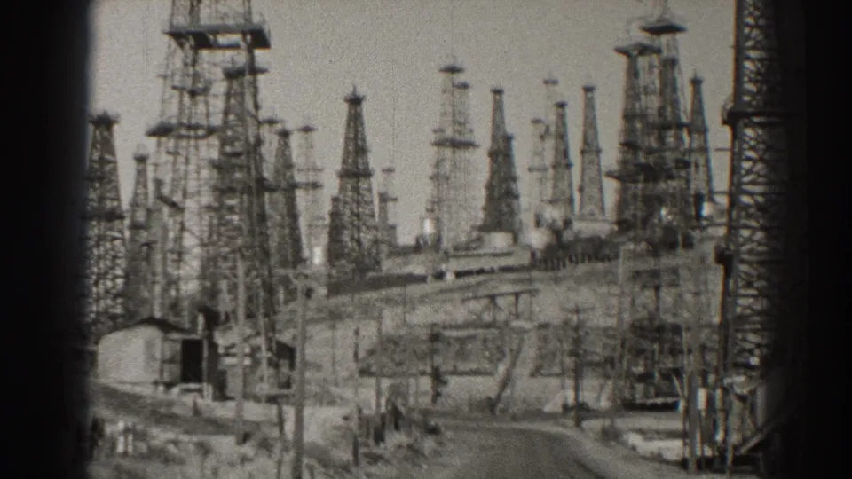 1937 Long Beach 8mm frame showing a road through a power-station yard with lattice towers, tanks, poles, and industrial equipment.