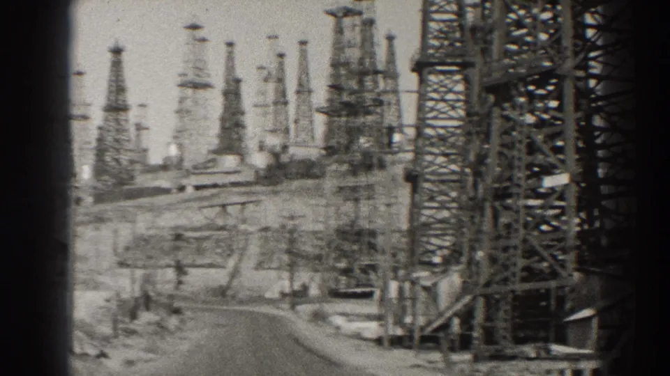 1937 Long Beach oil-field frame showing derricks, service road, and industrial structures through 8mm grain.