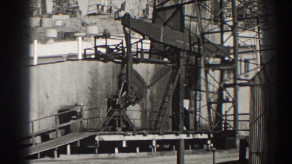 Close Long Beach oil-field view with a pumpjack, raised platform, steel tower, and large tank structure.