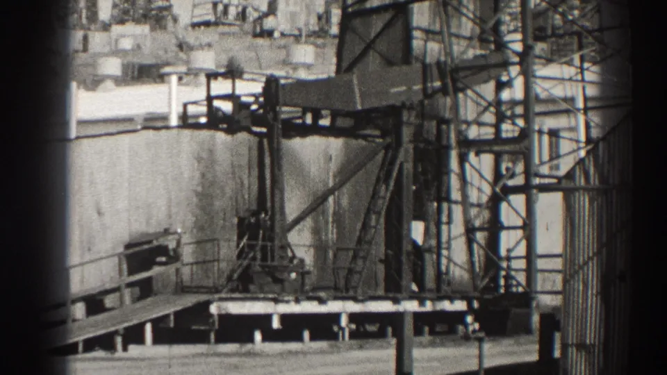 1937 Long Beach oil pumpjack scene with a pumping unit, platform railings, scaffolding, and tank structures in black-and-white 8mm footage.