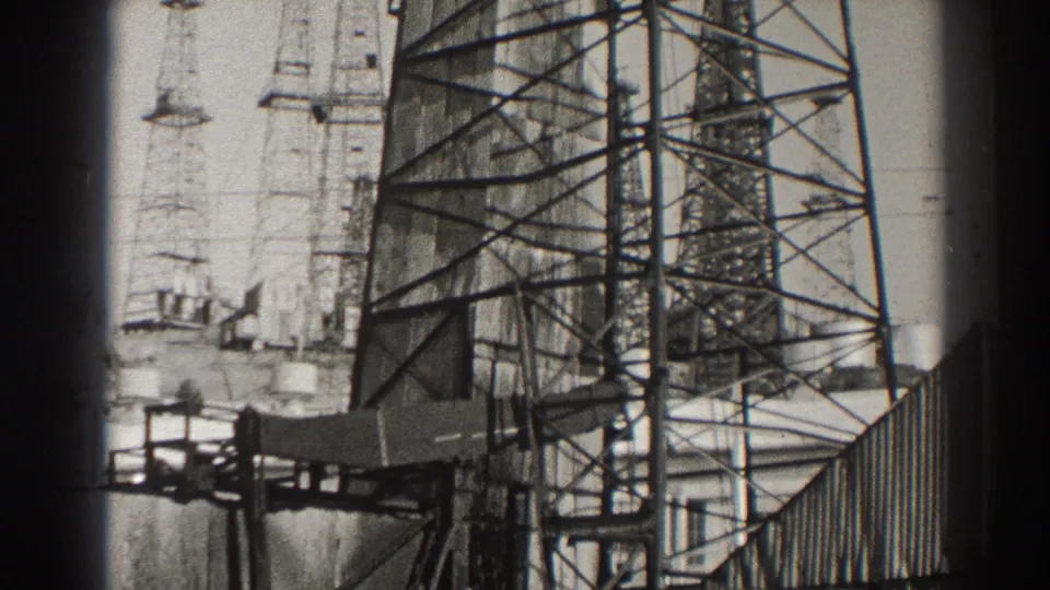 Black-and-white 8mm view of the Long Beach pumpjack at a different stroke position, surrounded by railings, tanks, and steel tower framing.