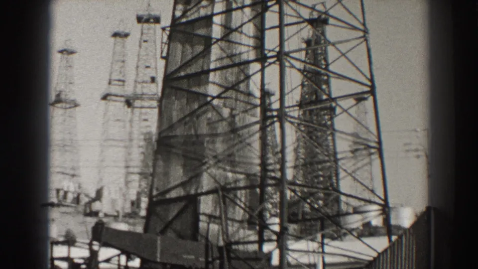 1937 Long Beach 8mm frame showing multiple tall oil derricks, with a large lattice tower in the foreground and smaller towers behind it.