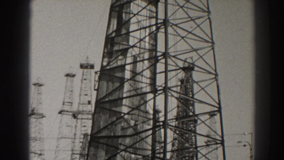 Black-and-white 1937 Long Beach frame with a prominent oil derrick at right and several smaller drilling towers across the skyline.