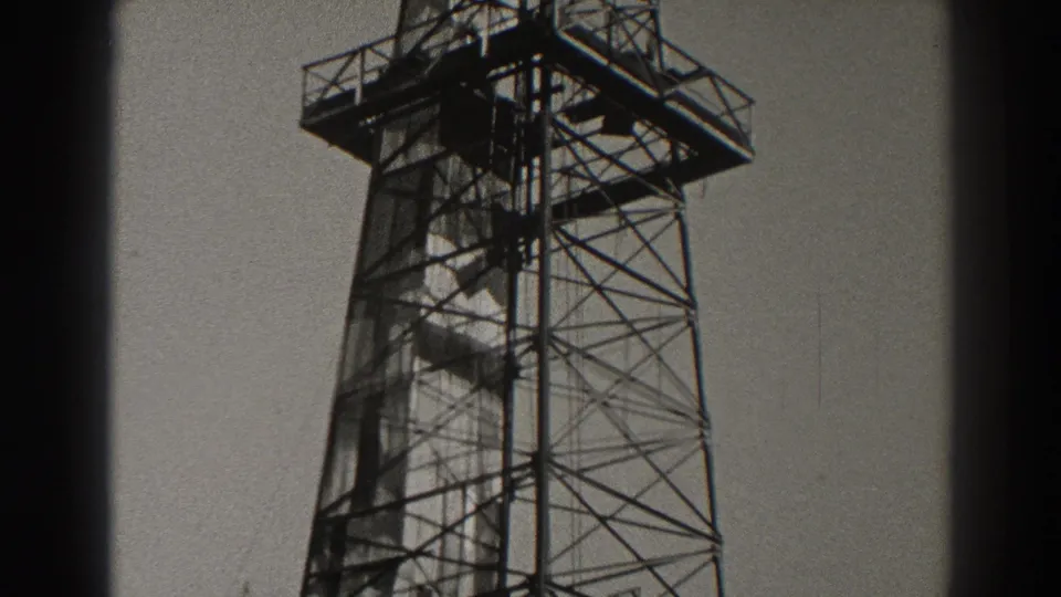 Black-and-white 1937 Long Beach frame showing an oil derrick with an elevated work platform, dense lattice bracing, and open sky behind it.