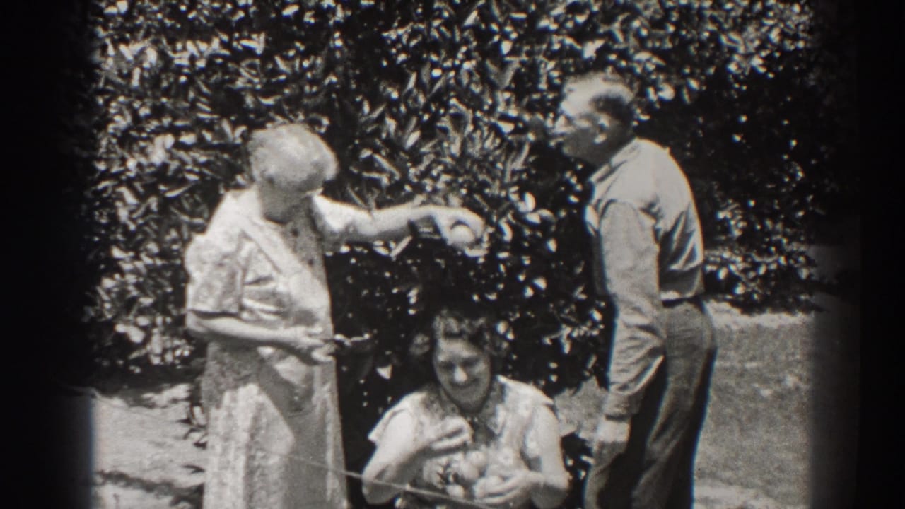 In a 1937 Long Beach garden scene, an older woman reaches toward fruit near a seated woman while a man stands beside them.