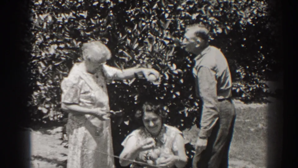 In a 1937 Long Beach garden scene, an older woman reaches toward fruit near a seated woman while a man stands beside them.