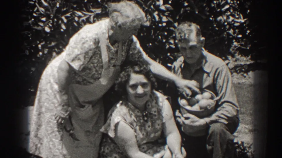An older woman in an apron leans toward a seated woman as a man holds a basket of fruit in 1937 Long Beach home-movie footage.