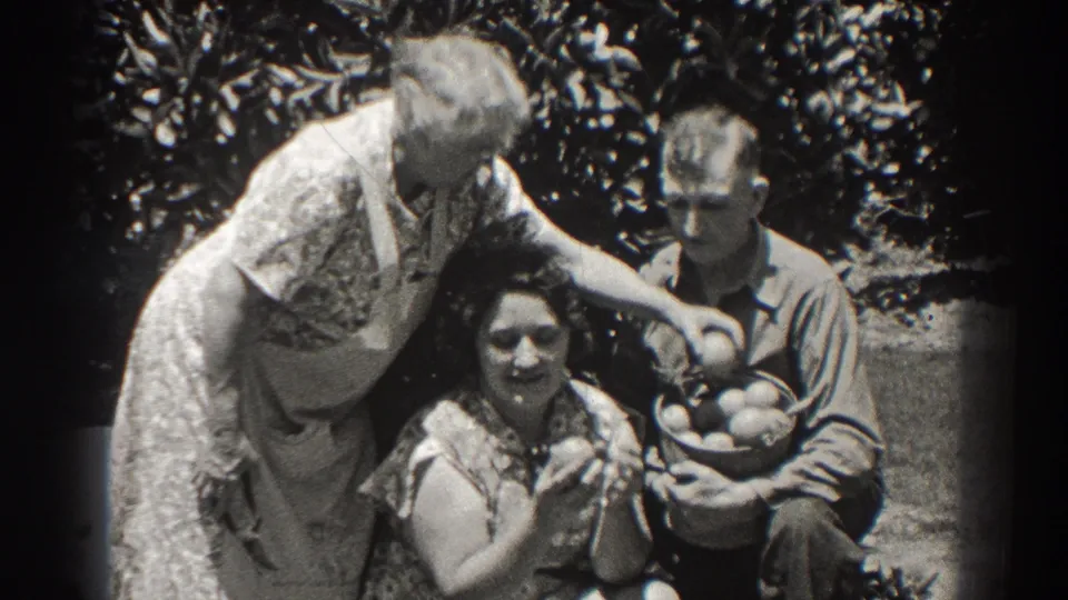 A seated woman lifts fruit toward the camera while an older woman reaches into a basket held by a man in the garden.