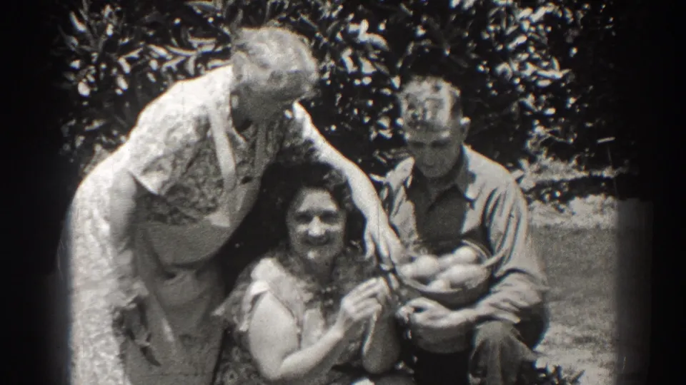 1937 Long Beach home-movie frame showing a woman in a white apron leaning over a seated woman while a man holds a basket outdoors.