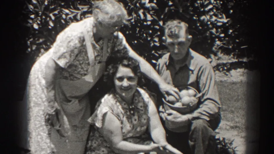 1937 Long Beach frame with three adults outdoors, including a standing woman in an apron reaching toward a basket held by a seated man.