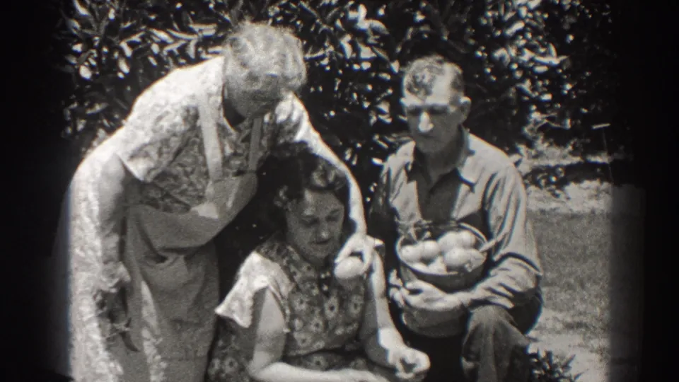 In this 1937 Long Beach outdoor home-movie frame, the apron-wearing woman bends close to the seated woman as the man holds the basket.