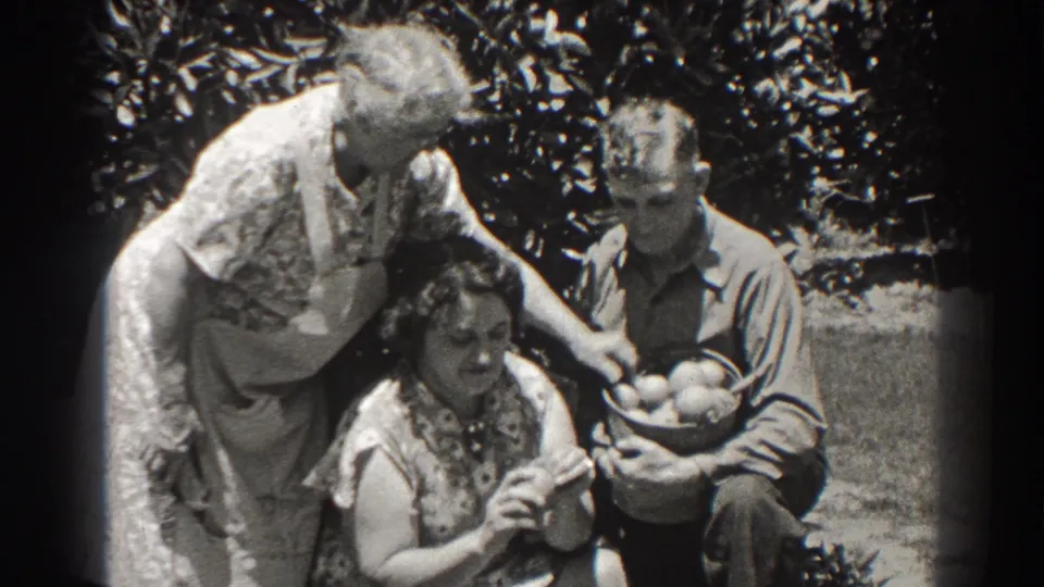1937 Long Beach outdoor frame showing a woman in an apron taking or selecting an object from a basket held by a seated man.