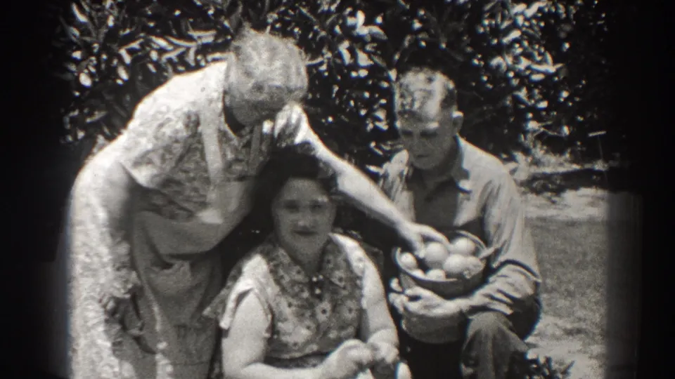 1937 Long Beach home-movie frame of three adults outdoors, with a woman leaning over another woman while a man holds a basket of fruit.