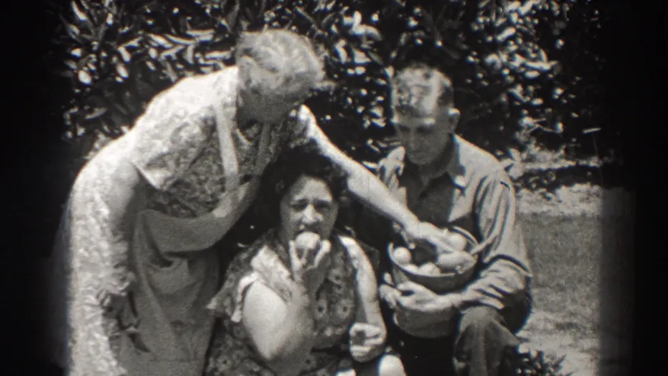 A seated woman appears to taste fruit in a 1937 Long Beach garden home movie, with another woman leaning nearby and a man holding more fruit.
