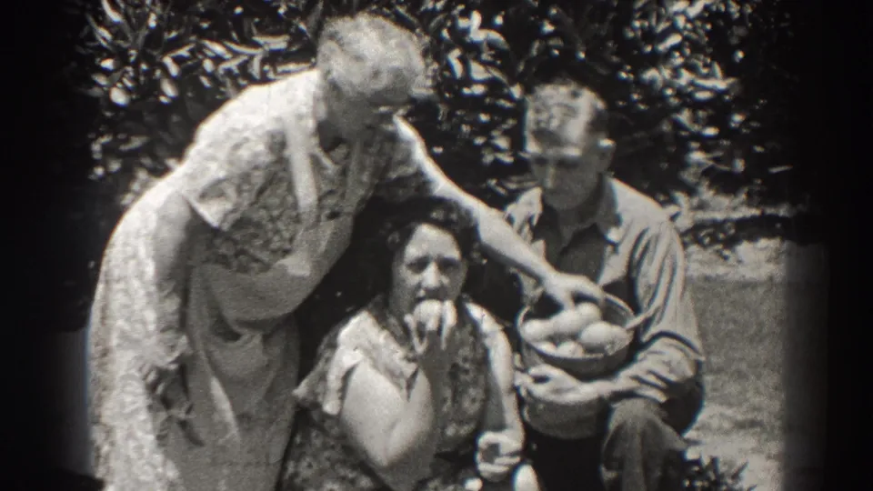 1930s home-movie frame of three adults outdoors, with one seated woman eating an apple and a man holding a basket of fruit.