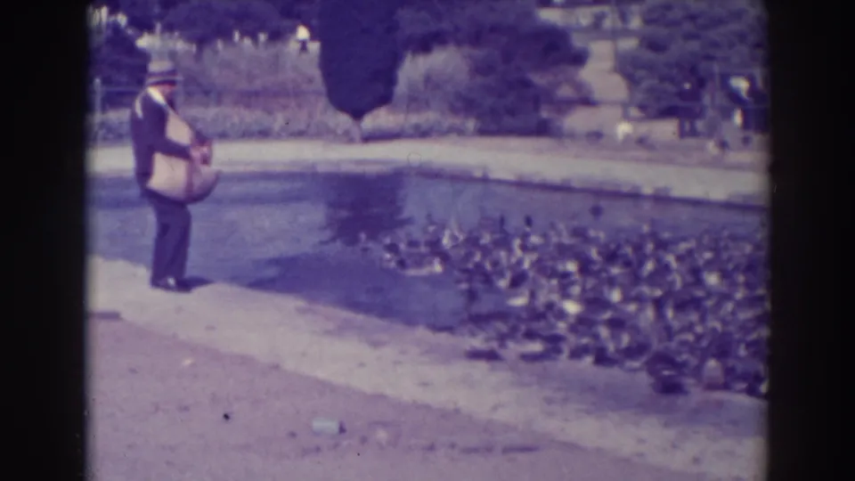 1937 Oakland home-movie frame showing a man with a bag feeding ducks gathered along a park pond.