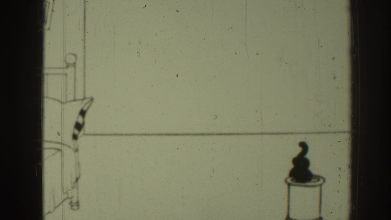 Small black cat sits on a bucket in a sparse cartoon room with a bed at left.