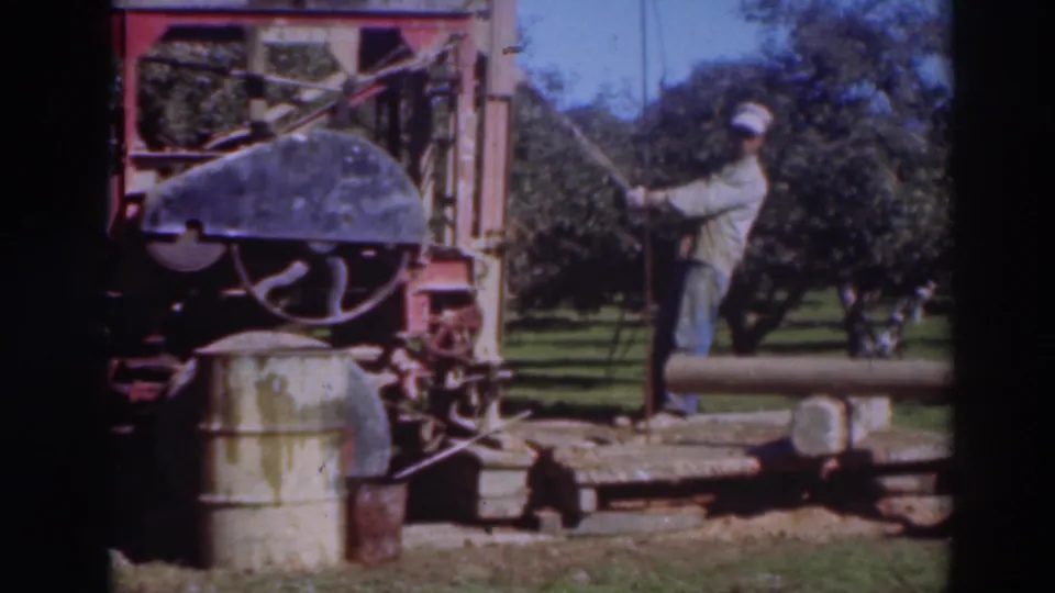1937 Sacramento 8mm frame showing a man operating or standing beside a large outdoor machine near orchard trees and a clear sky.