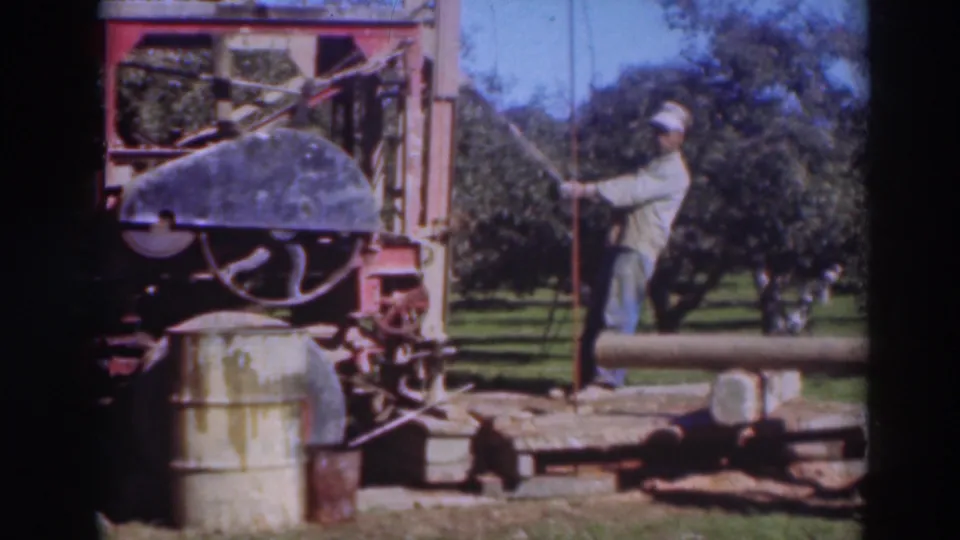 1937 Sacramento 8mm frame showing an outdoor machine, barrels, and a man holding a hose or pole in an orchard-like setting.