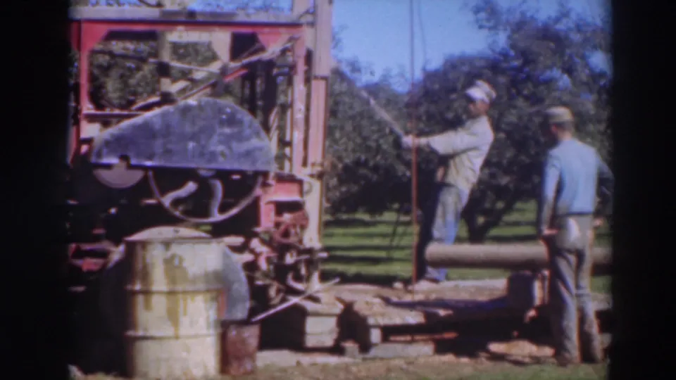 1937 Sacramento frame with one man operating the sawmill and another standing nearby beside a log in the orchard.