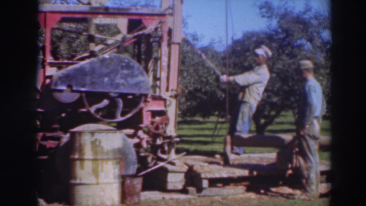Two adult men operate or observe a red drilling rig in a Sacramento orchard, 1937, with a barrel and trees visible.