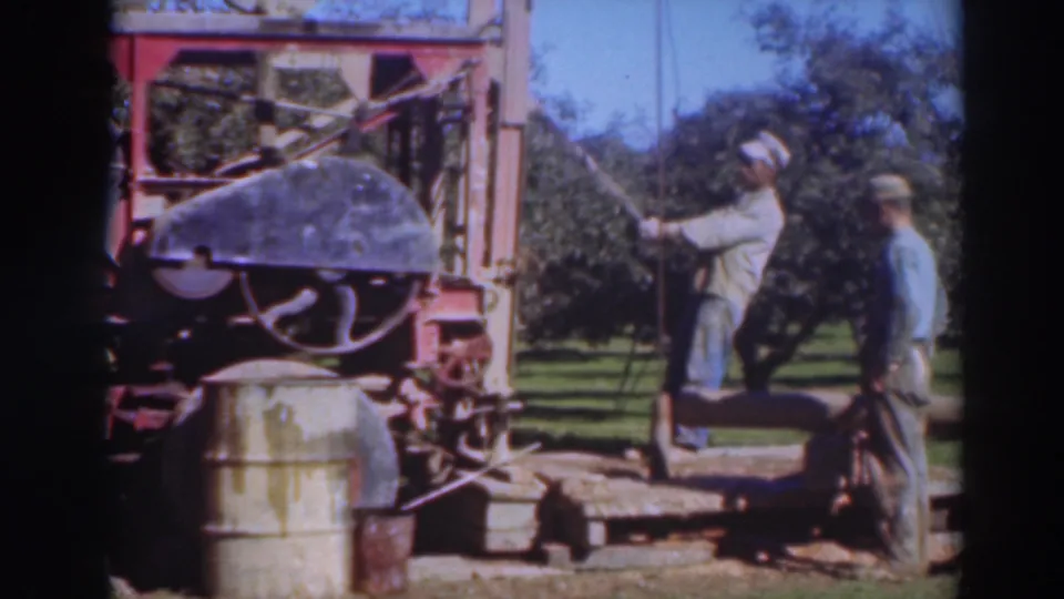 Two adult men operate or observe a red drilling rig in a Sacramento orchard, 1937, with a barrel and trees visible.