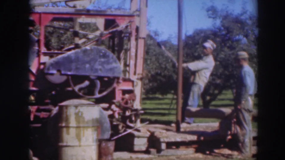 Two men remain beside a red drilling machine in the Sacramento orchard, with a vertical pole and barrel in the foreground.