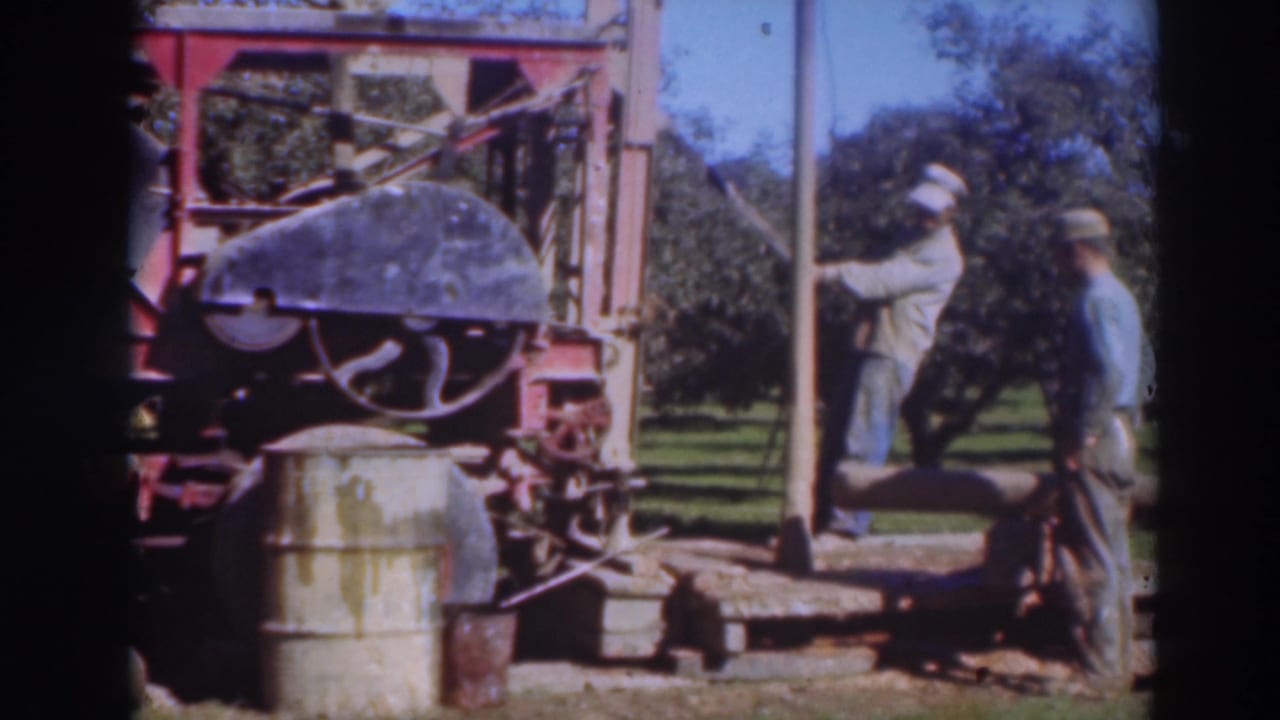 Two men work near a red rig and metal barrel in an orchard.