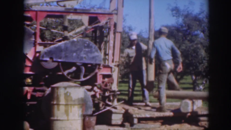 Two male workers in caps stand on a low wooden platform beside the red rig, with orchard trees behind them.