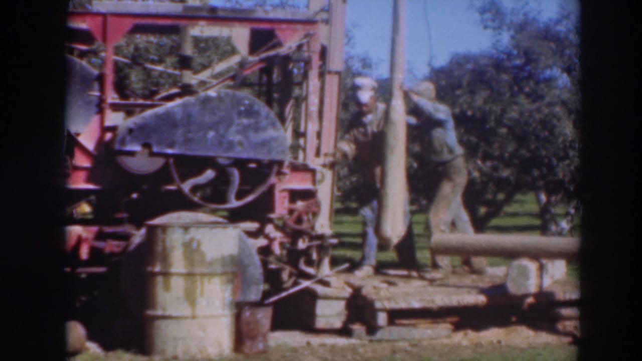 In 1937 Sacramento home-movie footage, two adult men handle a long wooden pole beside a red industrial machine in a tree-lined outdoor setting.