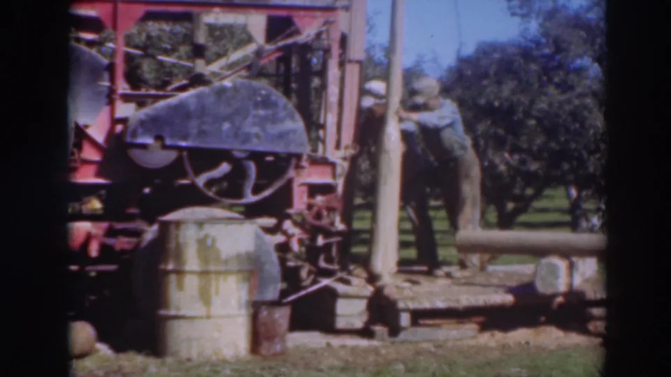 A worker in period clothing stands near the upright wooden pole and red machine, with trees and grass visible in the Sacramento outdoor setting.