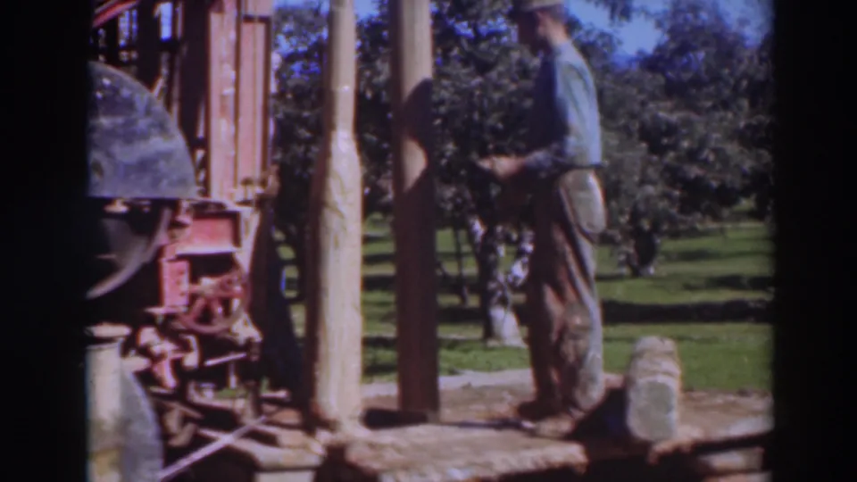 The 8mm frame shows a worker close to a vertical wooden pole, with the red machine at left and orchard-like trees in the background.