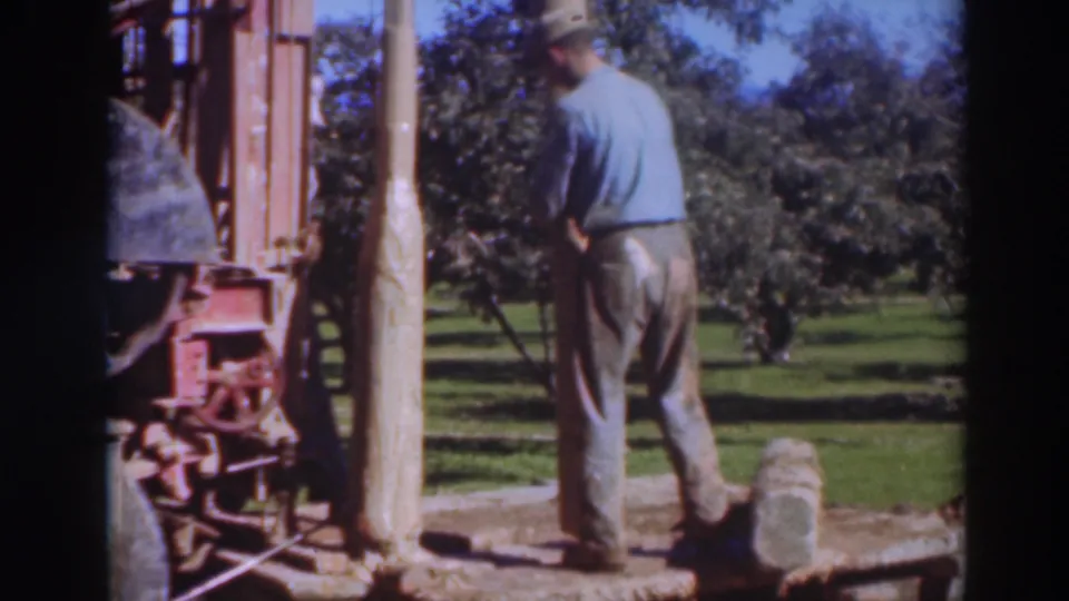 A worker seen from behind handles the area around the wooden pole and red machine, with open grass and trees beyond the work site.