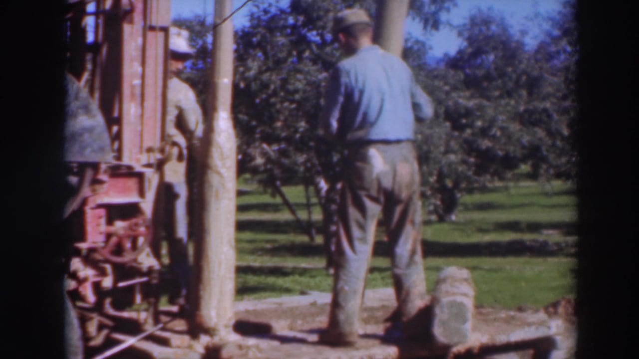 In 1937 Sacramento home-movie footage, two adult men stand near orchard rows while handling a long wooden beam at a small outdoor worksite.