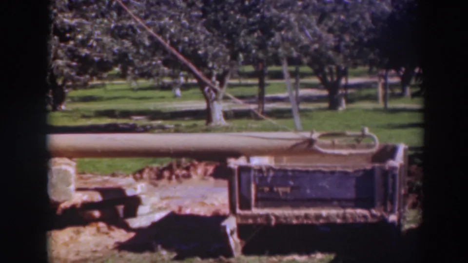 1937 Sacramento home-movie frame with a rope tied near a long wooden beam, a metal cart-like frame, and orchard-like trees beyond.