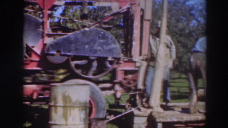 1937 Sacramento 8mm frame showing two adult men in work clothes near a red industrial machine, drum, hose, grass, and trees.