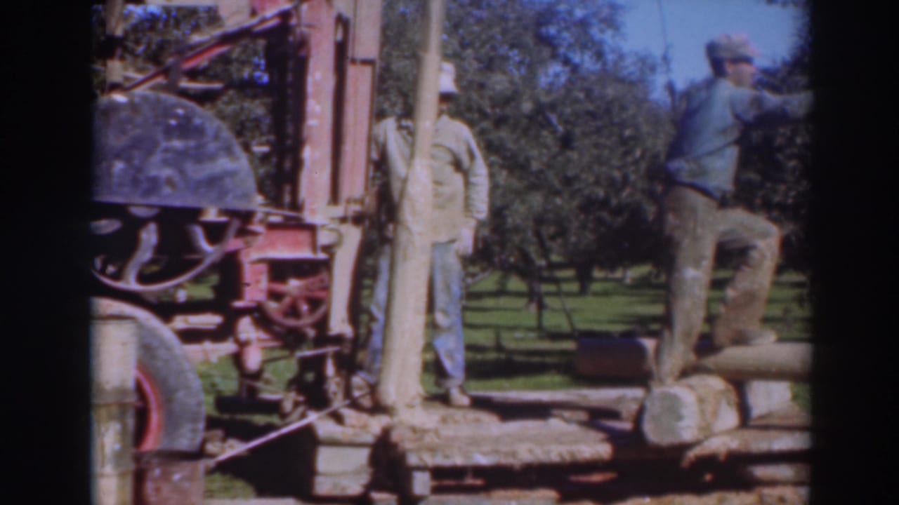Two men stand beside a red portable saw rig and long logs in a grassy tree-lined setting.