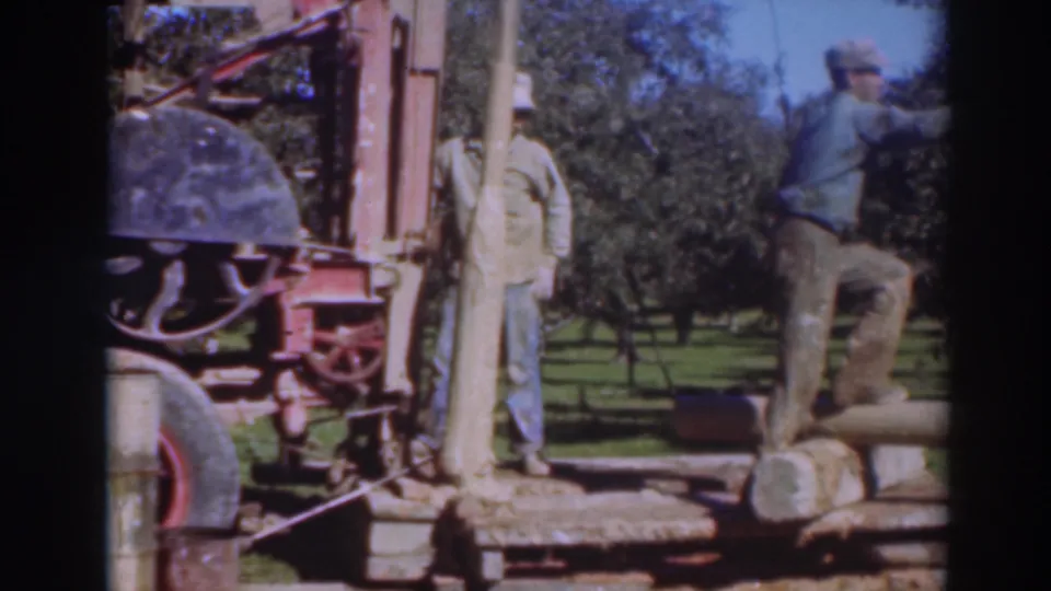 1937 Sacramento-area home-movie frame showing two adult men working near a red saw rig, long timber, grass, and trees.