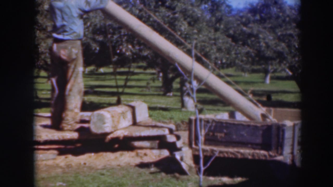 In 1937 Sacramento home-movie footage, two male workers stand beside a slanted lifting boom and cylindrical pipe section in an orchard.