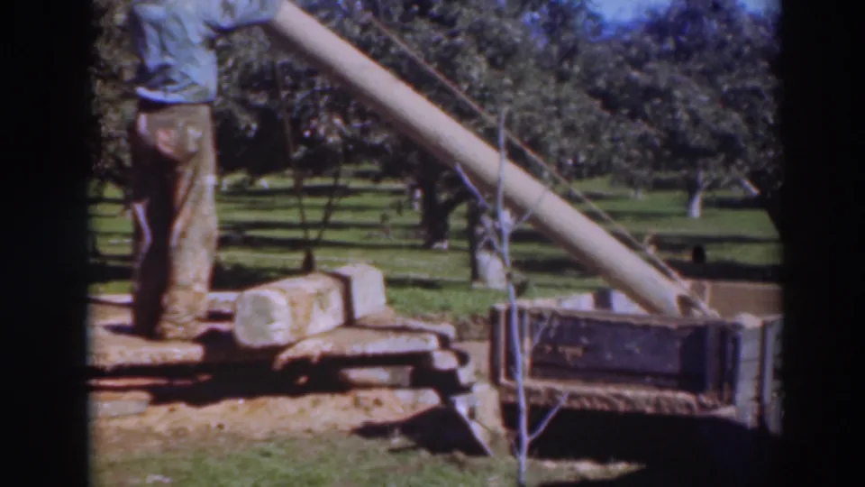 In 1937 Sacramento home-movie footage, two male workers stand beside a slanted lifting boom and cylindrical pipe section in an orchard.