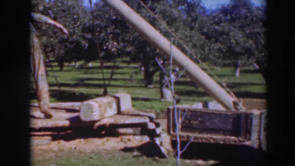 A male worker stands by the lifting rig and pipe section, with grass and mature orchard trees visible behind him.