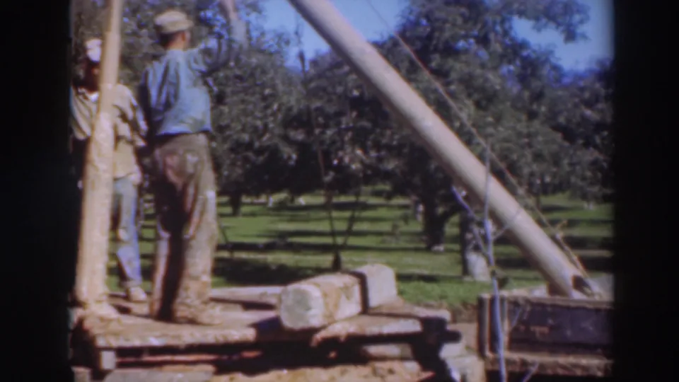 Two male workers in hats stand near the vertical support and pipe-lifting rig in the Sacramento orchard footage.